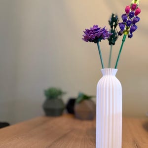 Könnte beinhalten: Eine weiße Keramikvase mit geriffeltem Design hält drei künstliche Blumen. Die Blumen sind lila, rosa und grün.