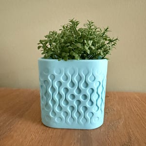 Op de afbeelding: Een lichtblauwe, vierkante bloempot met een herhalend golfpatroon, gevuld met groene bladplanten. De pot staat op een houten oppervlak, wat een contrast van kleuren en texturen creëert. Het ontwerp is modern en decoratief.
