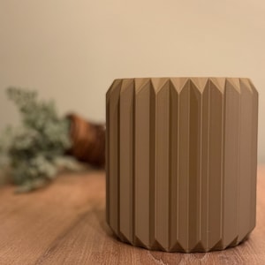Op de afbeelding: Een bruine, cilindrische plantenpot met een geometrisch, geribbeld ontwerp. De pot is gemaakt van een glad, mat materiaal.