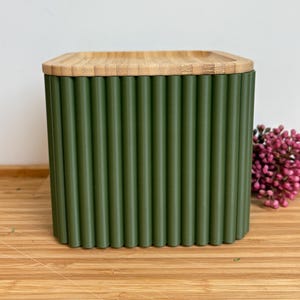 Könnte beinhalten: Ein quadratischer, olivgrüner Behälter mit vertikalen geriffelten Details. Der Deckel ist aus hellfarbenem Bambus gefertigt. Der Behälter steht auf einer Holzoberfläche, mit einer kleinen Gruppe rosa Blüten im Hintergrund.