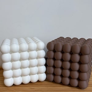 Könnte beinhalten: Zwei würfelförmige Dekorationsgegenstände. Einer ist weiß, der andere braun. Jeder Würfel besteht aus zahlreichen kugelförmigen Formen, die ein strukturiertes, blasenartiges Aussehen erzeugen. Die Objekte stehen auf einer Holzoberfläche.