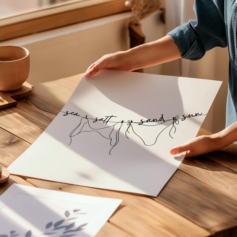 May include: A white art print featuring a minimalist line drawing of a bikini and the words "sea, salt & sand & sun" in black script. The print is held by a person, with a wooden table and a cup in the background.