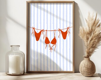 May include: A framed art print featuring a minimalist illustration of a red bikini hanging on a clothesline. The background has blue and white vertical stripes. The artwork is displayed on a wooden surface with a vase and candle holder.