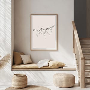 May include: A framed art print with a minimalist line drawing of three bikini tops hanging on a line. The words "sea, salt, sand, sun" are written above the drawing in a delicate script. The artwork is displayed in a light-colored room.