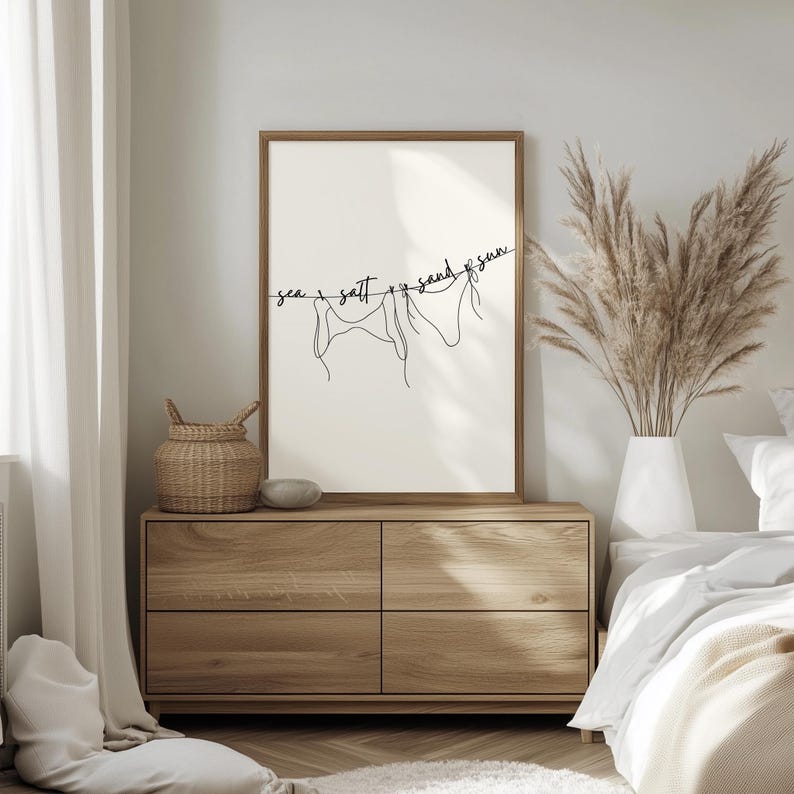 May include: A framed art print featuring a minimalist line drawing of three swimsuits hanging on a clothesline. The words "sea, salt, sand, sun" are written above the swimsuits in a delicate script. The artwork is displayed on a wooden dresser.