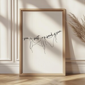 May include: A framed art print featuring a minimalist line drawing of three figures hanging on a clothesline. The words "sea, salt, sand, sun" are written above the figures in a cursive font. The frame is a light brown color.