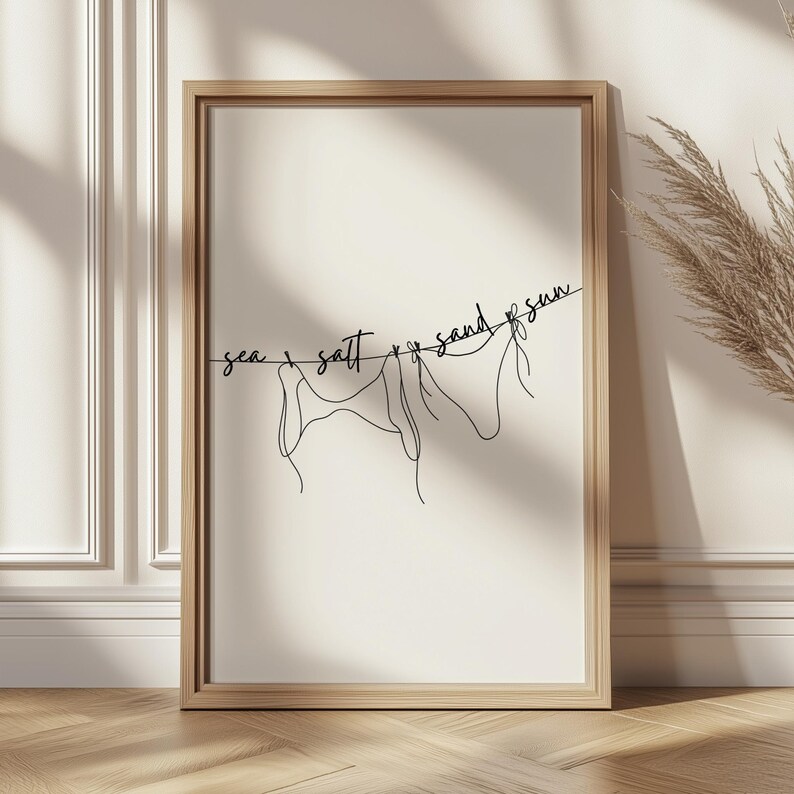 May include: A framed art print featuring a minimalist line drawing of three figures hanging on a clothesline. The words "sea, salt, sand, sun" are written above the figures in a cursive font. The frame is a light brown color.
