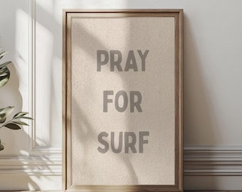 May include: A framed wall art print with the words "PRAY FOR SURF" in gray letters. The artwork has a neutral color palette and a wooden frame. The print is displayed against a white wall and wooden floor.