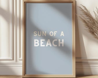 May include: A framed art print with the words "SUN OF A BEACH" in white letters on a light blue background. The frame is made of light brown wood. The print is leaning against a white wall and a wooden floor.