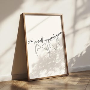 May include: A framed print featuring a minimalist line drawing of swimwear hanging on a clothesline. The words "sea, salt, sand, sun" are written above the swimwear in a cursive font. The frame is made of light brown wood.