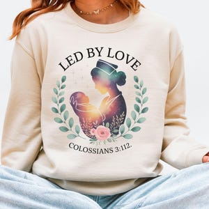 May include: A beige sweatshirt featuring the text "LED BY LOVE" above an illustration of a mother and child within a wreath. The illustration has a galaxy-like design. The text "COLOSSIANS 3:112" is below the illustration. The person in the image is wearing light blue jeans and white sneakers.