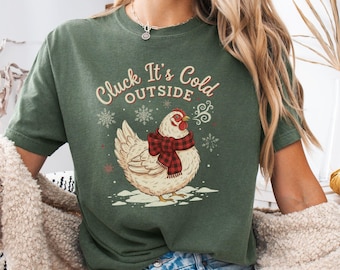 May include: Green t-shirt with the phrase "Cluck It's Cold Outside" and a graphic of a chicken wearing a red and black plaid scarf. The chicken is standing in snow with snowflakes around it.