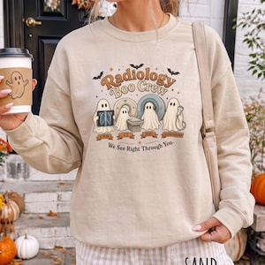 May include: A sand-colored crewneck sweatshirt featuring a graphic print with the text Radiology Boo Crew and We See Right Through You. The design includes four ghosts interacting with medical imaging equipment labeled X-ray, CT, MRI, and Ultrasound.