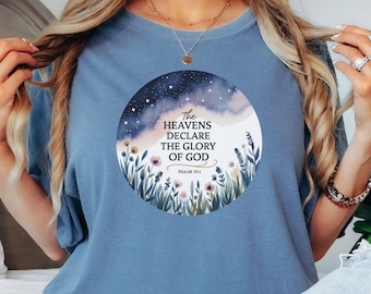 May include: A blue t-shirt featuring a circular graphic with the text "The Heavens Declare The Glory of God" and Psalm 19:1. The graphic includes a watercolor design of a starry night sky and flowers.