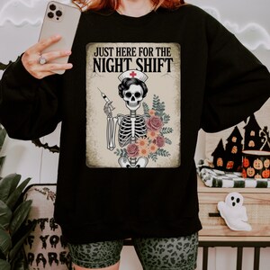 May include: Black sweatshirt featuring a graphic of a skeleton nurse holding a syringe and flowers, with the text "JUST HERE FOR THE NIGHT SHIFT". The design is on a beige background.