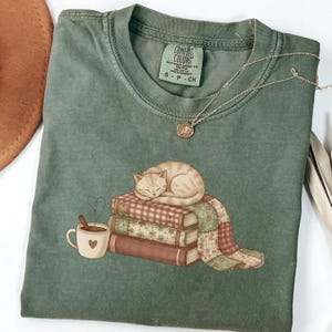 May include: Sage green Comfort Colors t-shirt featuring a graphic of a sleeping cat atop a stack of books, a cup of coffee, and a heart-shaped steam. A silver necklace with a pendant rests on the shirt.