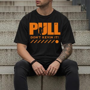 Puede incluir: Camiseta negra con la palabra "PULL" en naranja, una silueta de una persona apuntando con una escopeta y el texto "DON'T KEVIN IT!". Debajo, líneas y un círculo naranja. Una prenda de vestir informal.