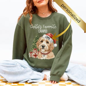 May include: Green sweatshirt with a graphic of a Cavapoo wearing a Santa hat. The text reads "Santa's Favorite Cavapoo" above the dog. A Christmas tree and wrapped gifts are also featured in the design.