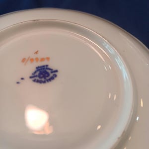 Peut inclure: Le dessous d'une soucoupe en c&eacute;ramique blanche avec un motif bleu et or. La soucoupe porte le num&eacute;ro "0/0007" imprim&eacute; en orange au fond.