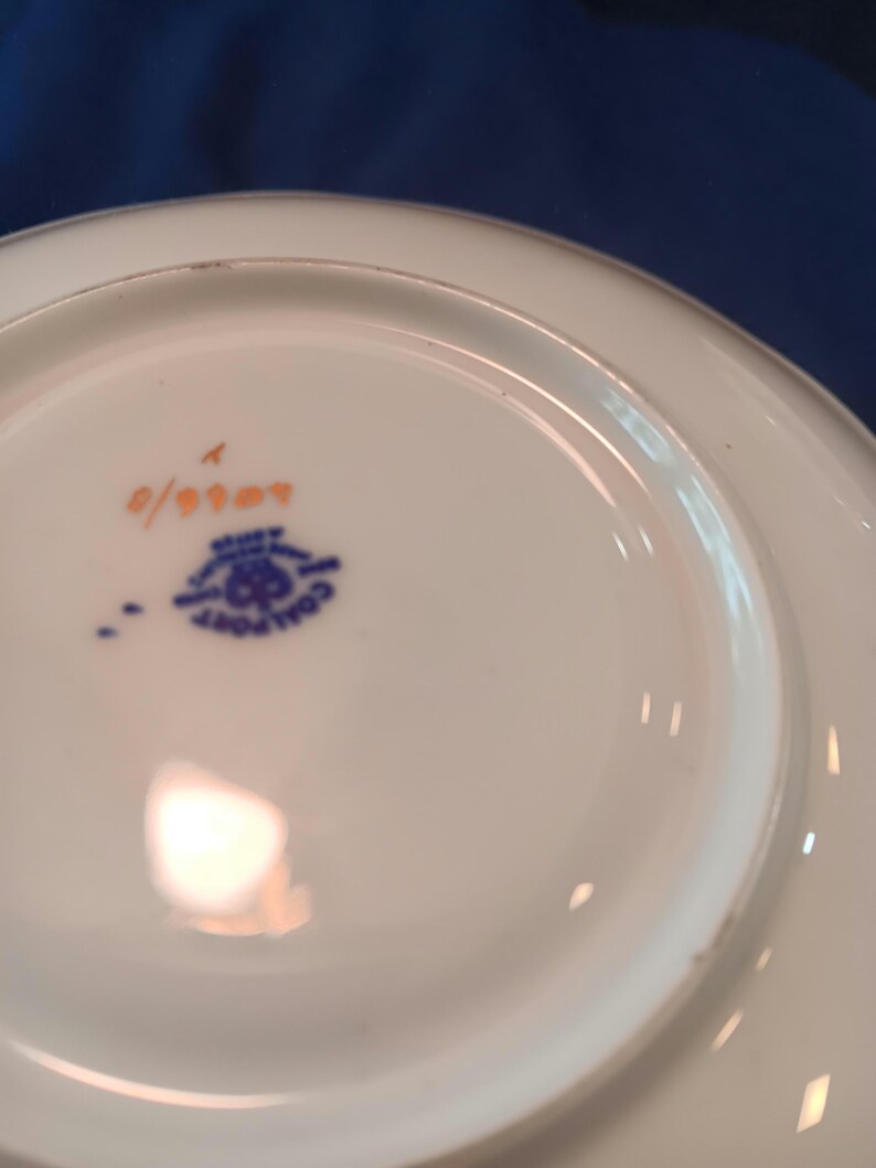 Peut inclure: Le dessous d'une soucoupe en c&eacute;ramique blanche avec un motif bleu et or. La soucoupe porte le num&eacute;ro "0/0007" imprim&eacute; en orange au fond.