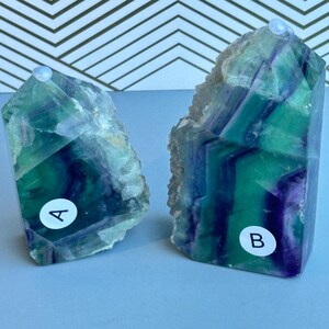 May include: Two polished fluorite crystal towers featuring vibrant bands of green and deep purple. The smaller tower is labeled with a white circular sticker marked A, and the larger tower features a white circular sticker marked B.