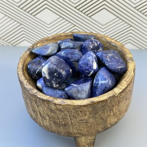 May include: A wooden bowl with three legs holds a collection of smooth, blue and white, tumbled Sodalite stones.