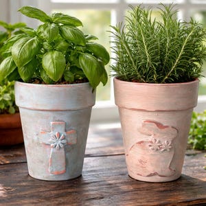 以下が含まれることがあります： 2つのテラコッタ製の装飾的な植木鉢。1つは十字架のデザインのライトブルー、もう1つはウサギのデザインのピンクです。どちらの鉢も、植物が入った状態で、窓の前の木製のテーブルの上に置かれています。