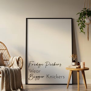 May include: A black framed poster with the text "Fridge Pickers Wear Bigger Knickers" hanging on a beige wall. A wicker chair with a beige throw blanket is in the foreground. A small wooden side table with a book and candles is in the background.