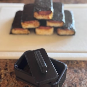 May include: A black plastic mold with a lid, designed for making rectangular shaped food items. The mold is open and the lid is sitting next to it.