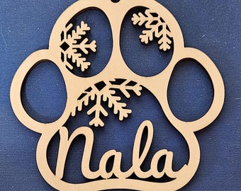 Könnte beinhalten: Ein Holz-Anhänger in Form einer Pfote mit Schneeflocken und dem ausgeschnittenen Namen "Nala". Der Anhänger ist hellbraun und hat oben ein Loch zum Aufhängen.