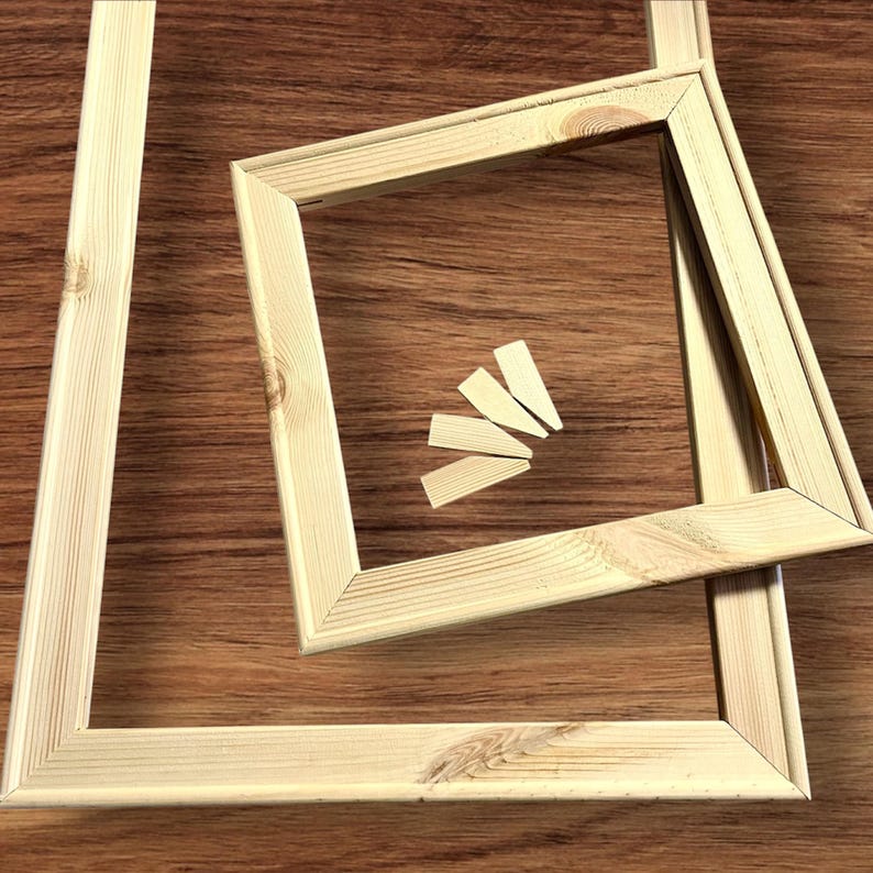 Two light-colored wooden stretcher bar frames, one large and one small, are arranged on a dark wood-grain surface, with four small wooden wedges in the center.
