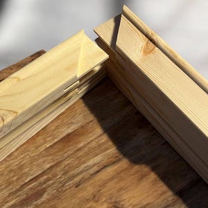 A close-up shows two light-colored wooden stretcher bar pieces joined at a corner, resting on a dark wood surface.