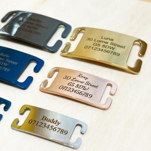 May include: A collection of rectangular metal pet ID tags in various metallic finishes including gold, rose gold, silver, blue, and dark grey. Each tag features side slots for collar attachment and is engraved with a name, address, and phone number.