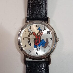 May include: A silver wristwatch with a black leather band features a white face with black numbers and a cartoon image of Goofy. The watch face also includes the text "Goofy" and "Disney."