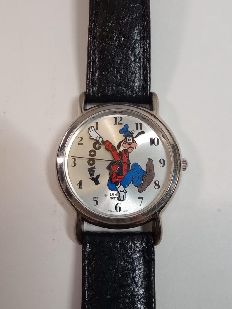 May include: A silver wristwatch with a black leather band features a white face with black numbers and a cartoon image of Goofy. The watch face also includes the text "Goofy" and "Disney."