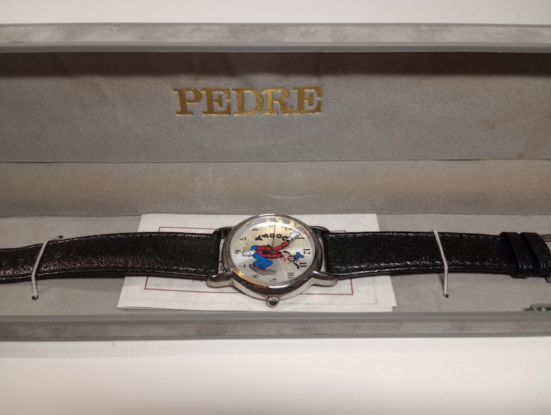 May include: A silver wristwatch with a black leather band. The watch face features a cartoon character with a red and blue outfit. The watch is in a gray box with the word "PEDRE" printed in gold letters.