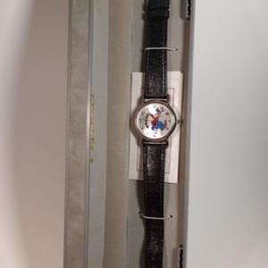May include: A silver watch with a black leather band and a white face featuring a cartoon image of Goofy. The watch is in a gray box.