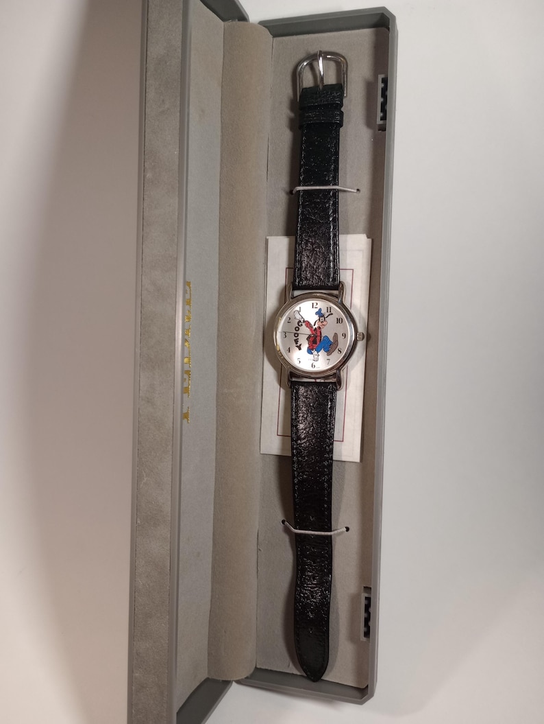 May include: A silver watch with a black leather band and a white face featuring a cartoon image of Goofy. The watch is in a gray box.