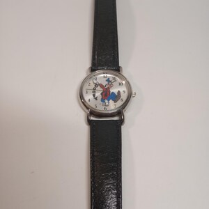 May include: A silver wristwatch with a black leather band. The watch face features a white background with a cartoon image of Goofy. The numbers 1 through 12 are printed around the edge of the watch face.