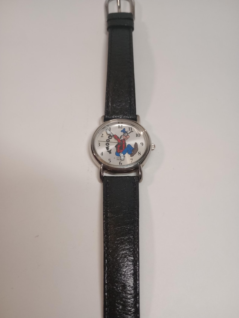 May include: A silver wristwatch with a black leather band. The watch face features a white background with a cartoon image of Goofy. The numbers 1 through 12 are printed around the edge of the watch face.