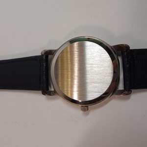 May include: A silver wristwatch with a black leather band. The watch face is covered with a smooth, brushed metal back.