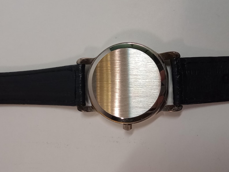 May include: A silver wristwatch with a black leather band. The watch face is covered with a smooth, brushed metal back.