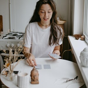 May include: A woman in a white t-shirt is arranging tarot cards on a white round table. A small Buddha statue, a lit candle, and a vase with dried flowers are on the table. A white Buddha head statue sits on a windowsill.