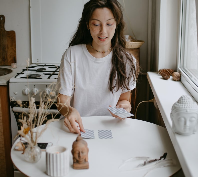 May include: A woman in a white t-shirt is arranging tarot cards on a white round table. A small Buddha statue, a lit candle, and a vase with dried flowers are on the table. A white Buddha head statue sits on a windowsill.