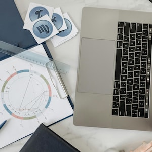May include: A white sheet of paper with a circle chart and a ruler on top of a blue folder. A laptop computer with a black keyboard is open on a white surface. There are several stickers with astrological signs on the paper.
