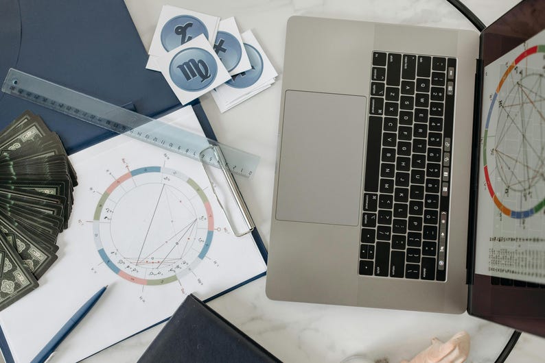 May include: A white sheet of paper with a circle chart and a ruler on top of a blue folder. A laptop computer with a black keyboard is open on a white surface. There are several stickers with astrological signs on the paper.