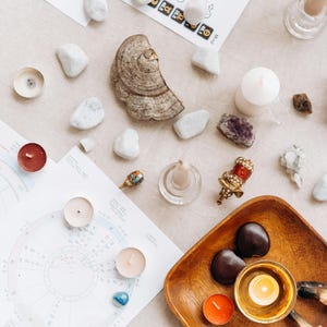 May include: A flat lay of various items used in a spiritual or meditation practice, including candles, crystals, stones, and a wooden bowl with a candle inside. The items are arranged on a light beige surface.