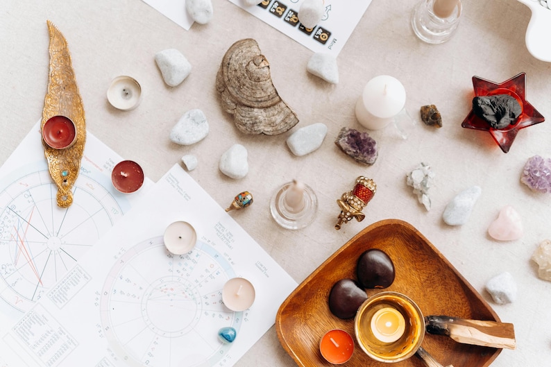 May include: A flat lay of various items used in a spiritual or meditation practice, including candles, crystals, stones, and a wooden bowl with a candle inside. The items are arranged on a light beige surface.