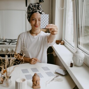 May include: A person holding a tarot card in front of their face. The person is wearing a white t-shirt and a patterned headscarf. A round white table holds several tarot cards, a candle, and a small Buddha statue. The scene is set in a well-lit room with a window.