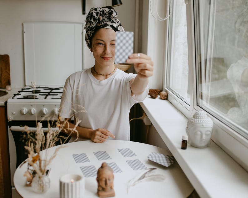 May include: A person holding a tarot card in front of their face. The person is wearing a white t-shirt and a patterned headscarf. A round white table holds several tarot cards, a candle, and a small Buddha statue. The scene is set in a well-lit room with a window.